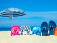 Beach summer holiday banner background. Flip-flops and hat with a board and ball on the sand near the ocean. Summer accessories on the seashore. Tropical vacation and relax travel concept. Top view and copy space.